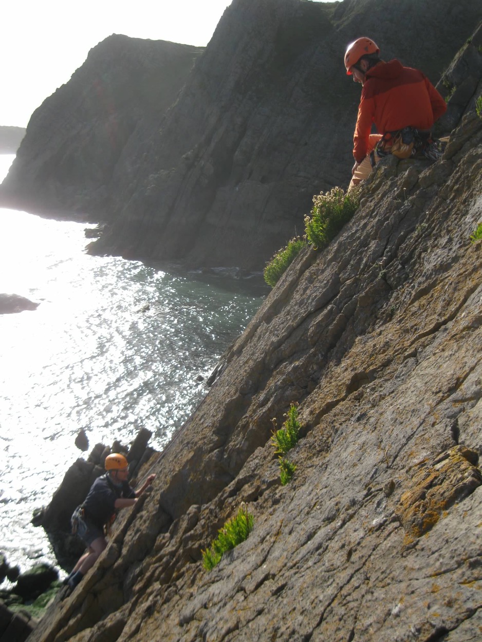 Phill sat at the top of a crag belaying a climber, with the sea in the background.